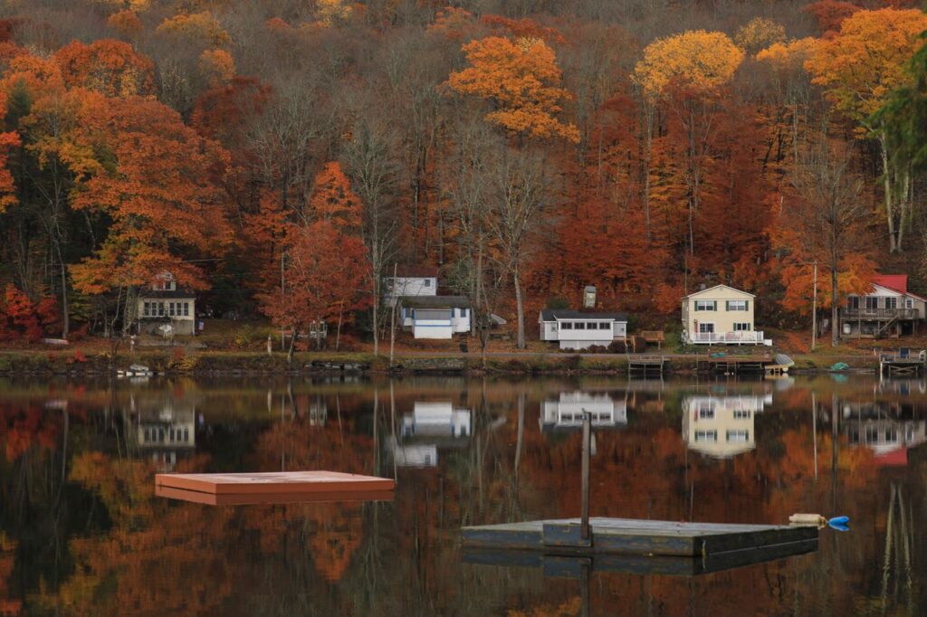 Autumn in the Catskills — road trip USA ideas Scenic view of autumn foliage reflecting on a calm lake with houses in the background. — road trip USA ideas