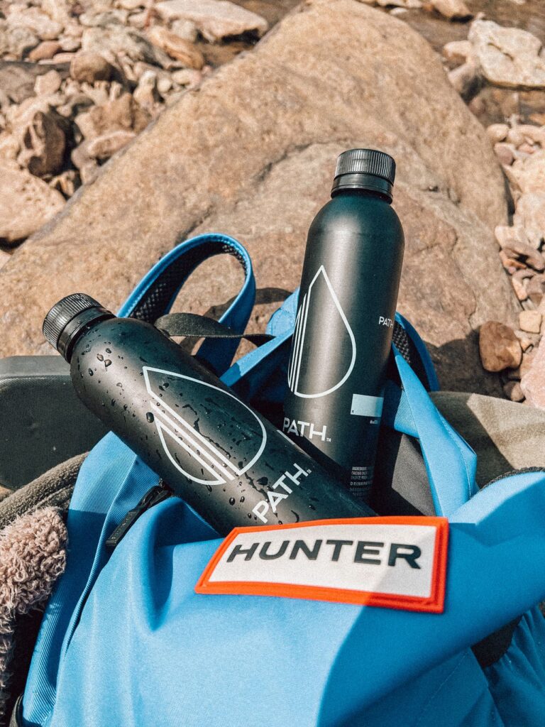 Reusable Water Bottles for Travel — travel essentials checklist Two reusable water bottles next to a blue backpack on rocky terrain. — travel essentials checklist